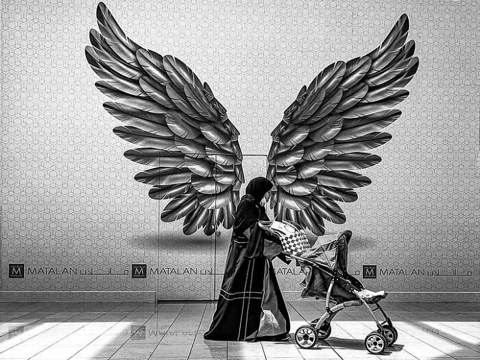 A woman pushing a stroller with large wings behind her, a striking photograph capturing life across cultures.