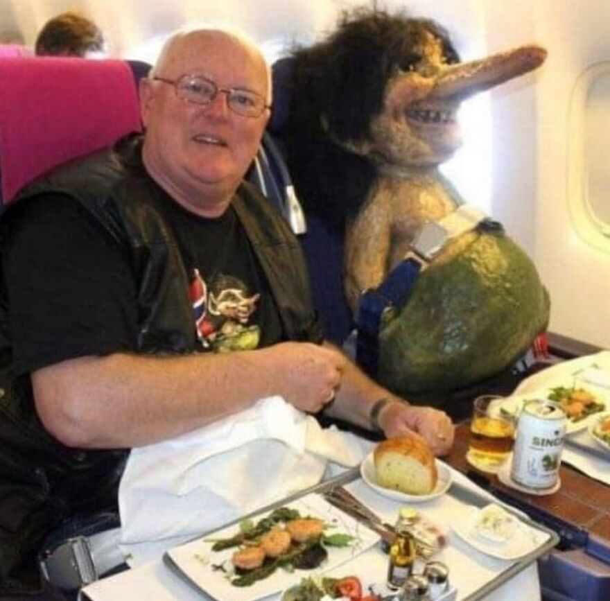 An unhinged image of a man and a creepy troll doll having a meal on an airplane that is truly cursed.