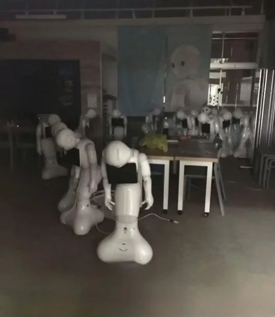 A dark room filled with several white humanoid robots standing idly, an unhinged and cursed image.