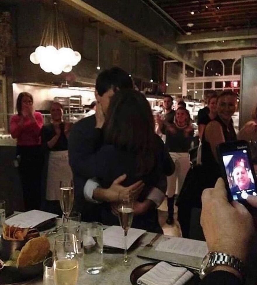 A cursed image of a couple hugging in a restaurant, surrounded by applauding people, while someone takes a photo with a phone.