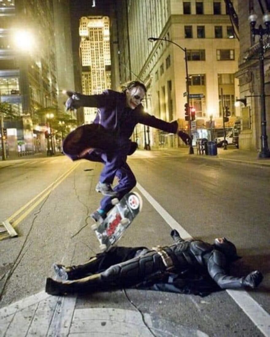 A cursed image showing Joker on a skateboard jumping over Batman lying on the ground in the middle of a city street at night.