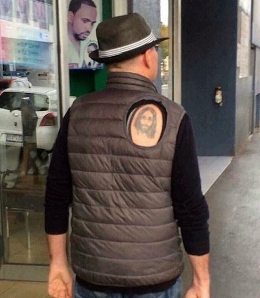 A man with a fedora and a vest showing a Jesus tattoo through a hole, a truly cursed image.