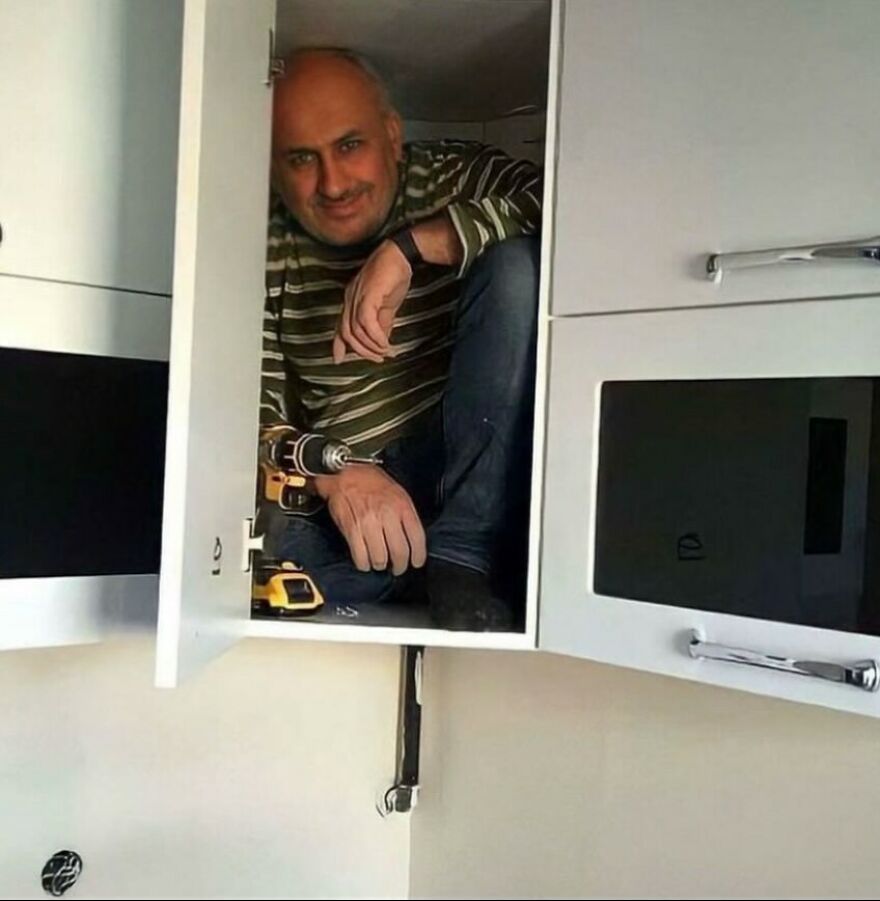 A man with a drill smiles from inside an overhead cabinet, a cursed image that is unhinged.