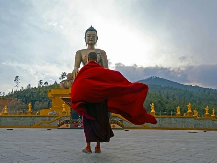 Remarkable photograph by Tittu Shaji Thomas of a monk in a red robe facing a large Buddha statue, capturing life across cultures and landscapes.
