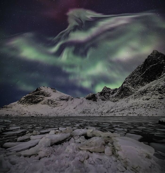 Jaw-dropping astronomy photo of vibrant green and purple aurora borealis over snowy mountains and a frozen lake.