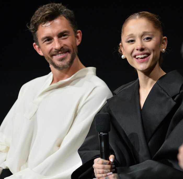 Ariana Grande smiling while holding a microphone, next to a man. She recently dropped out of an acting role. Ariana Grande smiling while holding a microphone, next to a man. She recently dropped out of an acting role.