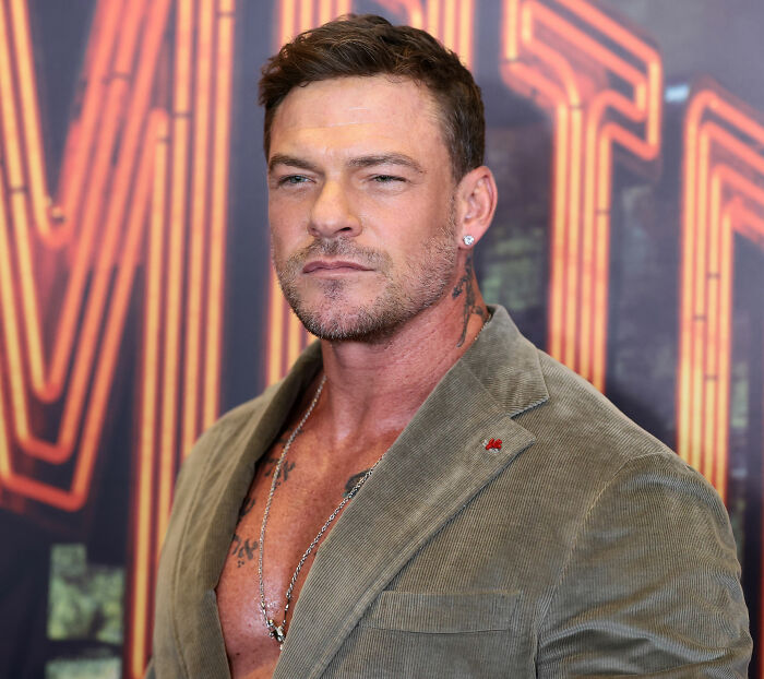 Alan Ritchson's shirtless moment at an event, revealing tattoos and a chain, with a serious expression. Alan Ritchson's shirtless moment at an event, revealing tattoos and a chain, with a serious expression.