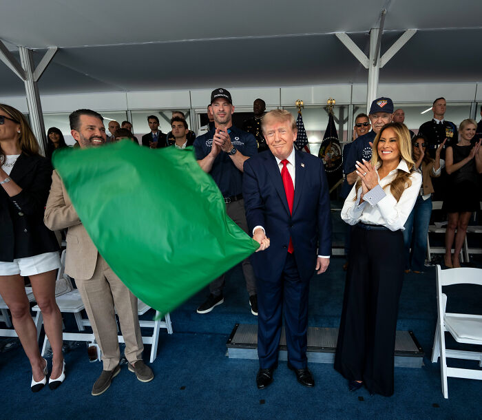 Donald Trump and Melania at IndyCar event, holding a green flag, with a heated exchange. Donald Trump and Melania at IndyCar event, holding a green flag, with a heated exchange.
