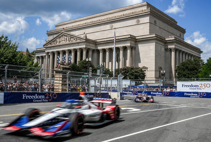 IndyCar event with racing cars speeding past a historic building, with crowds watching. IndyCar event with racing cars speeding past a historic building, with crowds watching.