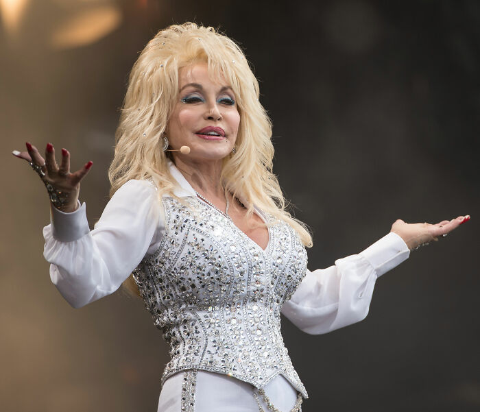 Dolly Parton on stage, performing in a glittering white outfit, discussing her illness and hospital admission.