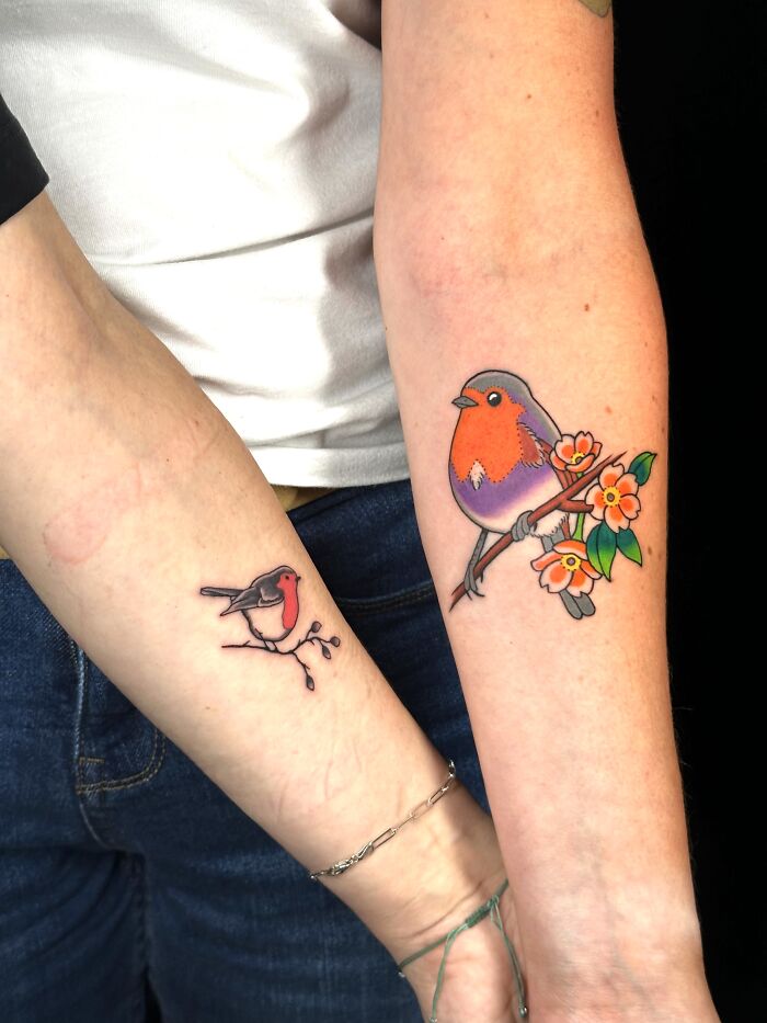 Two arms with matching ink of a small and large robin bird, perched on a branch with flowers, showcasing matching ink ideas.