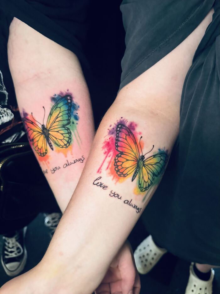 Two arms with vibrant, matching butterfly ink, each with 'love you always' text, showcasing matching ink ideas.