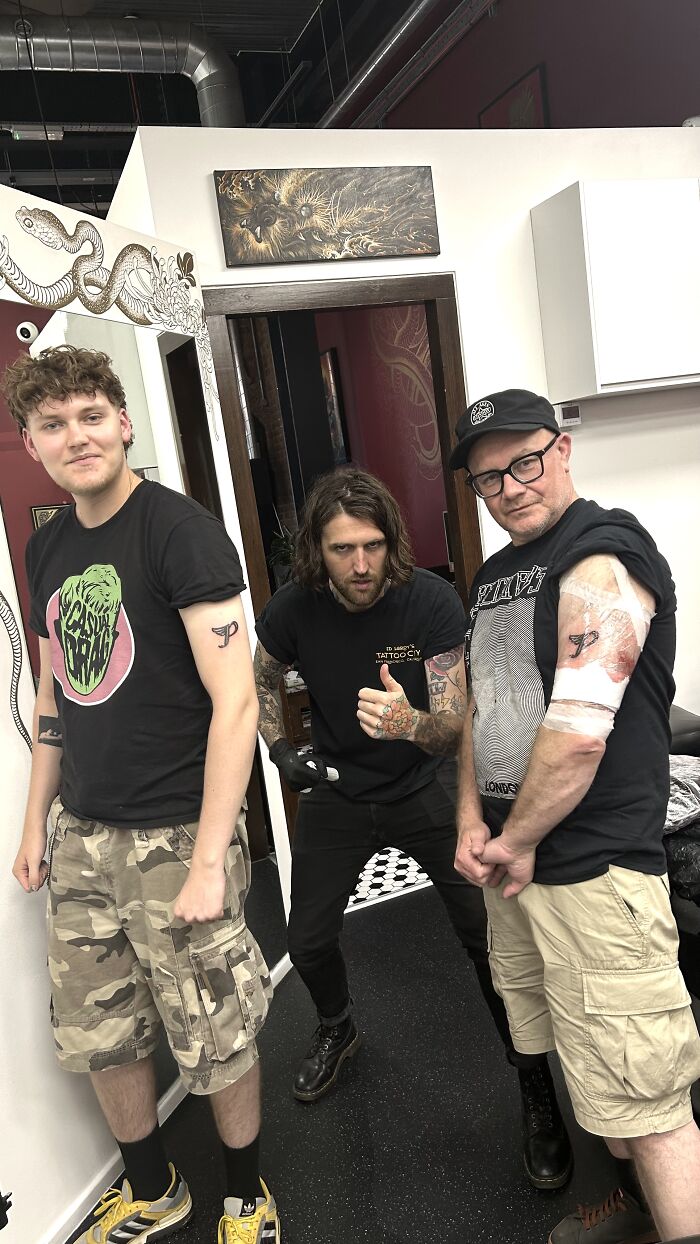 Three men pose in a tattoo studio, two of them showing off their fresh matching ink, demonstrating matching ink ideas.
