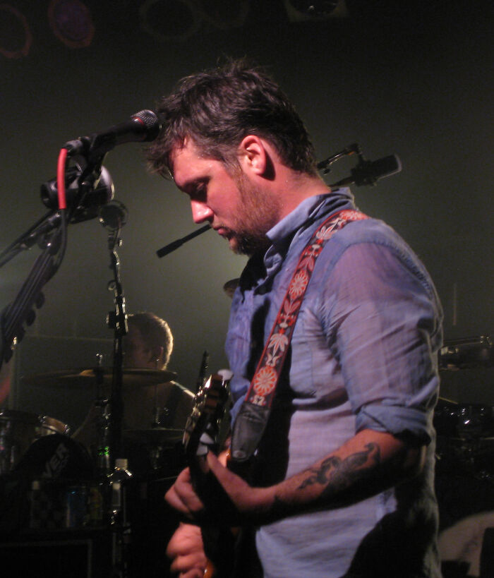 A male celebrity singer on stage, looking down while playing his guitar, illuminated by a spotlight, revealing what dating a celebrity is like.