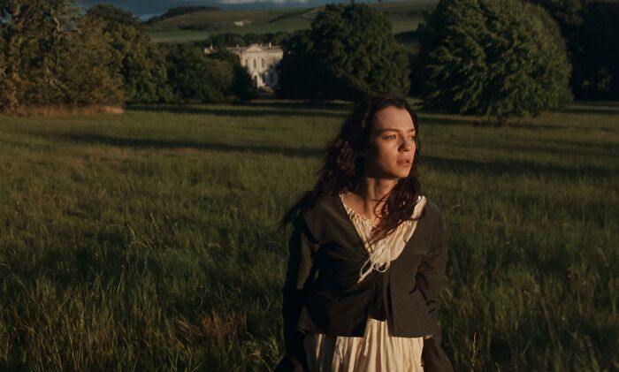 A woman in period clothing standing in a field with a mansion in the background, from a Jane Austen adaptation. A woman in period clothing standing in a field with a mansion in the background, from a Jane Austen adaptation.