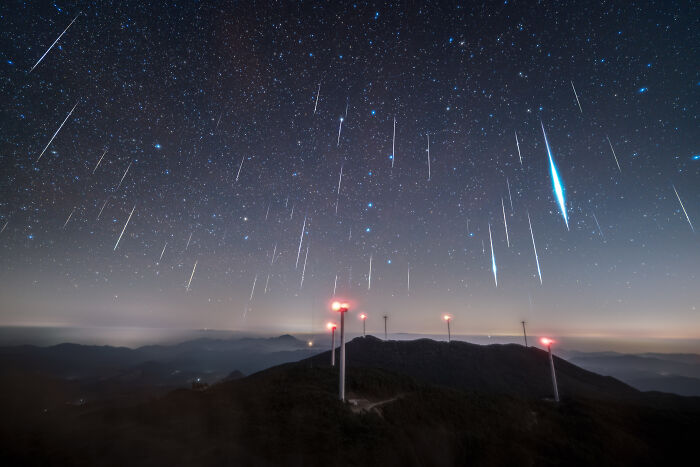 An incredible astronomy photo capturing a meteor shower streaking across the night sky above wind turbines on a mountain.