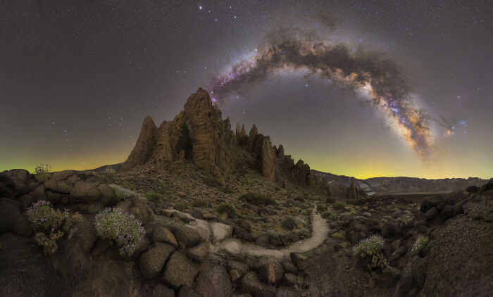 A breathtaking astronomy photo showing the Milky Way galaxy arching over rugged mountain peaks and a winding path.