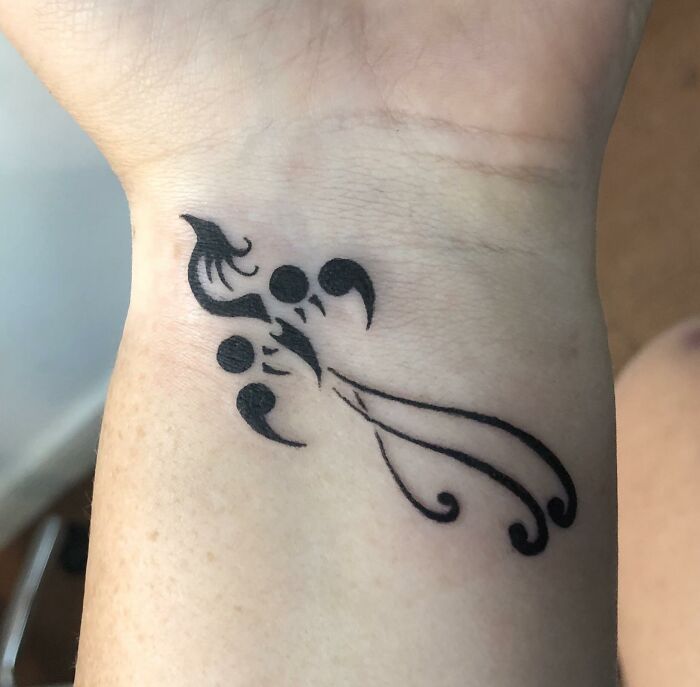 A black semicolon tattoo on a wrist, designed as a bird or phoenix with swirling lines, showing a powerful story.