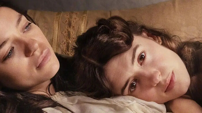 Two women with long hair lying down, one looking at the camera, in a scene from a Jane Austen adaptation. Two women with long hair lying down, one looking at the camera, in a scene from a Jane Austen adaptation.