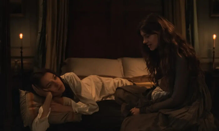 Two women in a dimly lit room, one sitting on a bed next to another who is lying down, from a Jane Austen adaptation. Two women in a dimly lit room, one sitting on a bed next to another who is lying down, from a Jane Austen adaptation.