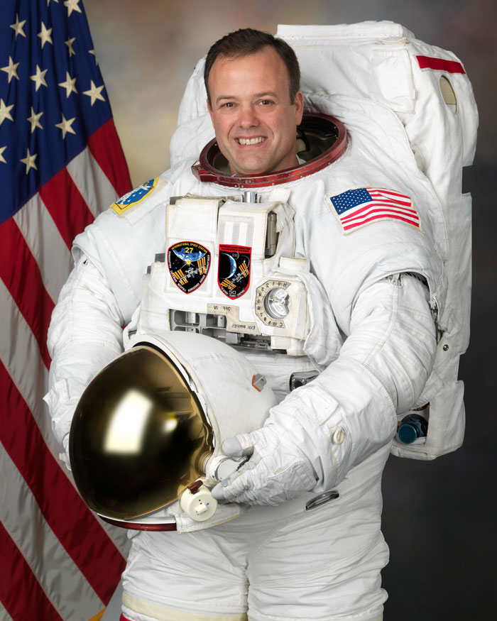 Astronaut smiling in a spacesuit, holding a helmet, against an American flag backdrop after 178 days in space.