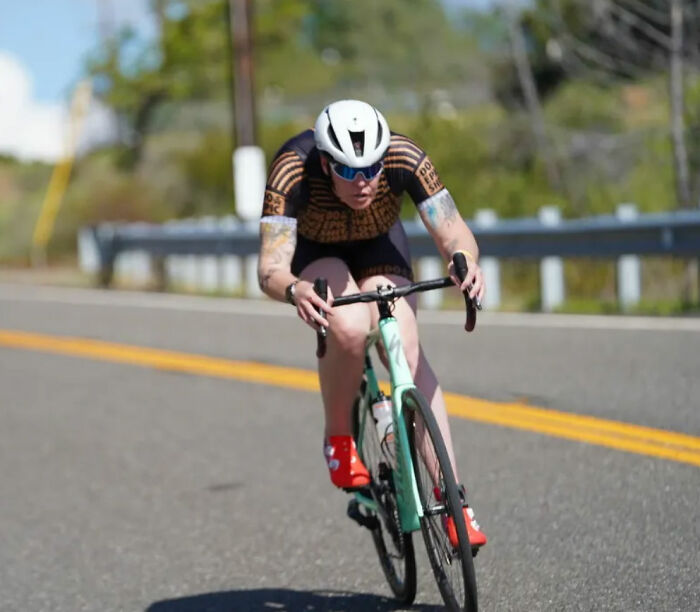 Trans American in a cycling suit and helmet, riding a bicycle on a road, a stark contrast to her European asylum experience. Trans American in a cycling suit and helmet, riding a bicycle on a road, a stark contrast to her European asylum experience.