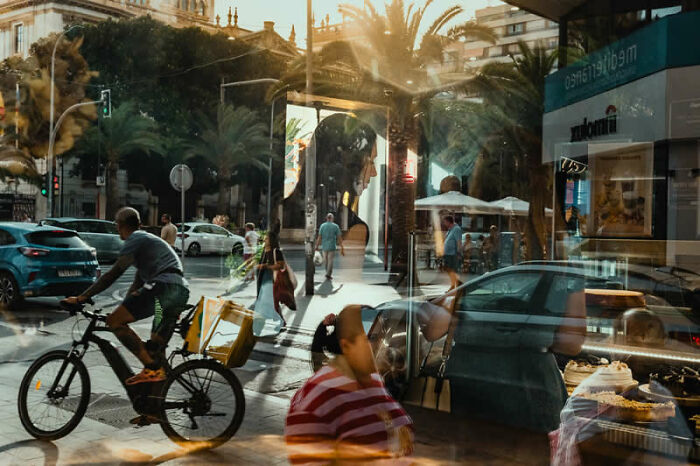 A street scene with reflections and a cyclist, showcasing urban life in a winning photo from LensCulture Street Photography Awards.