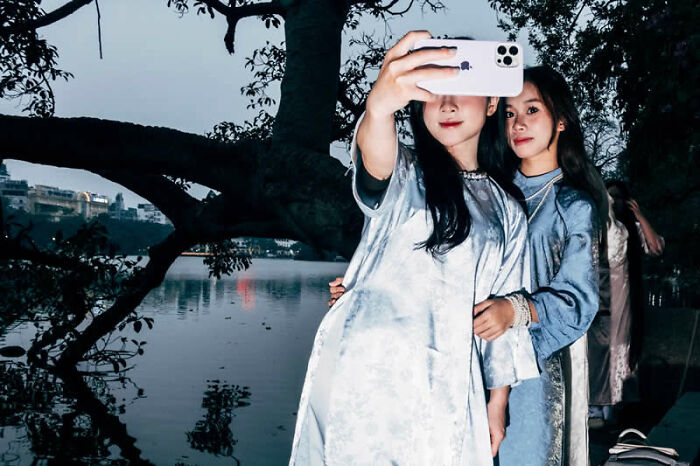 Two women in traditional dresses take a selfie by a lake, a winning photo from LensCulture Street Photography Awards.