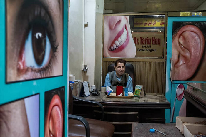 A man sitting at a desk in a cluttered room, surrounded by large posters of body parts, a winning street photography image.