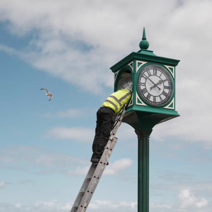 A worker on a ladder fixes a tall green clock tower under a cloudy sky, a striking image from the LensCulture Street Photography Awards.