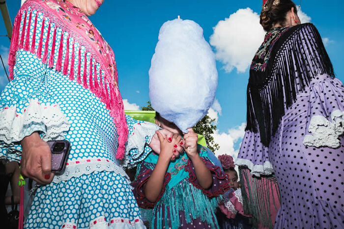 A girl in vibrant traditional attire eating cotton candy, surrounded by women in similar dresses at a street photography awards event.