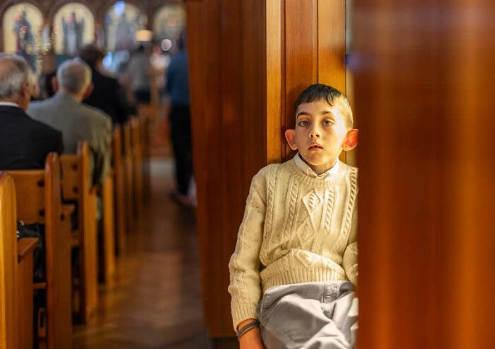 A young boy with prominent ears peers out from a wooden doorway during a church service, a winning photo from the LensCulture Street Photography Awards.