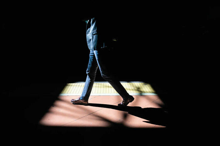 A person in jeans and a jacket walks through a spotlight on a dark ground, a captivating winning photo from the Street Photography Awards.