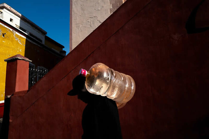 Person carrying a large water jug on their shoulder against a vibrant red and yellow building, a winning photo from the Street Photography Awards.