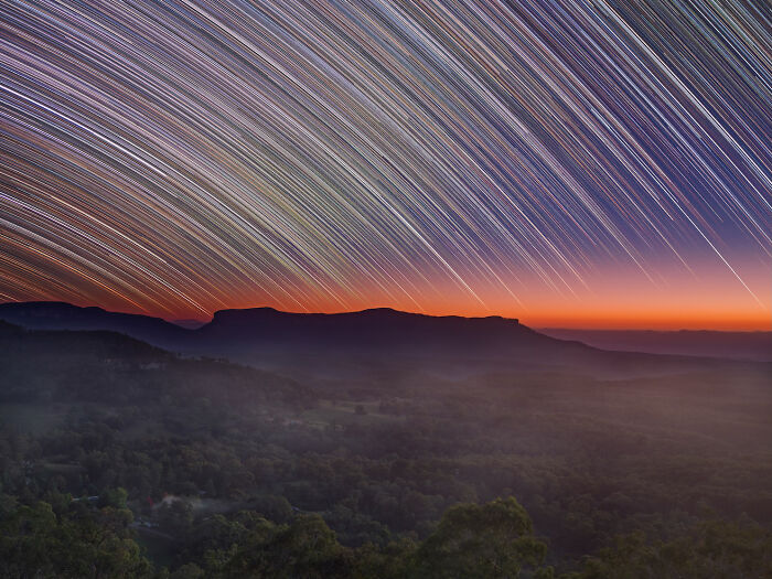 Astrophotography award winner: star trails arc across the sky over silhouetted mountains at sunrise.