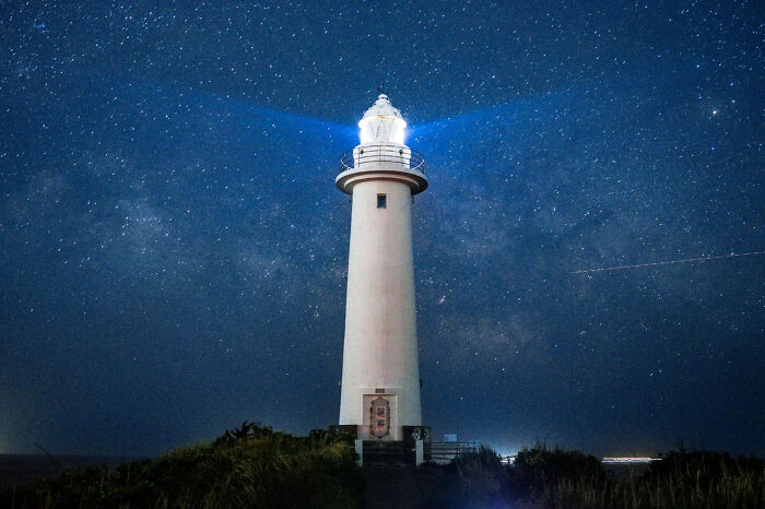 A brilliantly lit lighthouse stands against a dark, star-filled night sky, a winning image from the 2026 European Photography Awards.