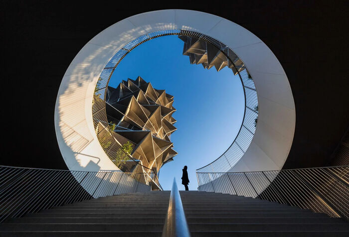 A person stands at the top of a modern circular staircase, looking up at an abstract building, an image from the 2026 European Photography Awards.