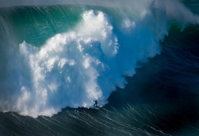A lone surfer on a small board rides a colossal blue and white ocean wave, one of the 2026 European Photography Awards winning images.