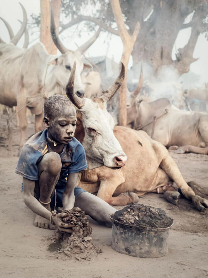 A young boy with a dirt-covered face crouches on the ground, creating shapes from mud. This image won a 2026 European Photography Award.