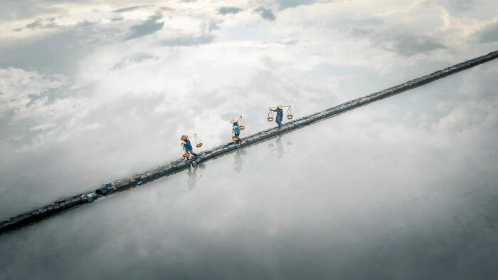 Three people in conical hats walk a narrow path above reflective water, a stunning image of the world from European Photography Awards.