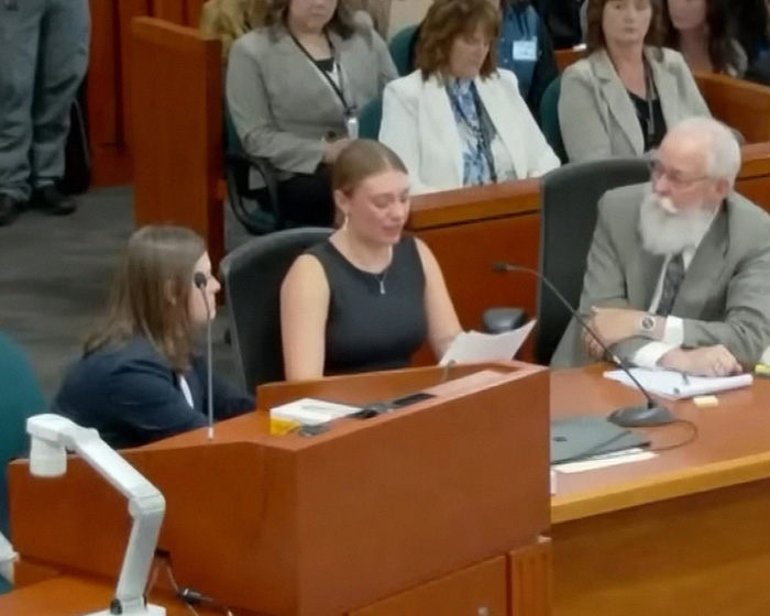 A woman in a black top reads from papers at a desk, with a man with a white beard next to her. Bryan Kohberger's case details.
