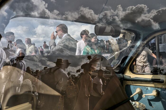 Reflection of clouds and people in a car window, showcasing authentic street photography moments.