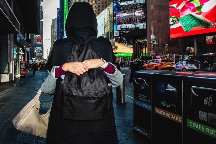 A person wearing a black coat and backpack walks through Times Square, capturing an authentic moment in street photography.