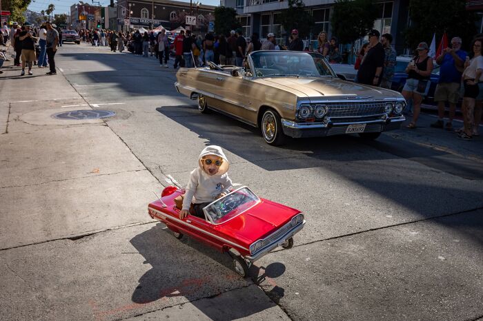 A young child in a red toy car with sunglasses, smiling, drives down a street near a classic car, an authentic street photography moment.