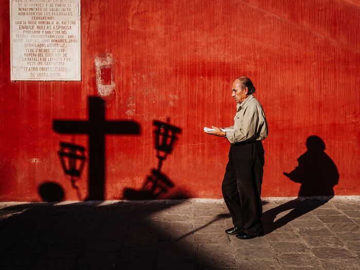 A man walks past a red wall, with strong shadows of a cross and lanterns, capturing an authentic street photography moment.