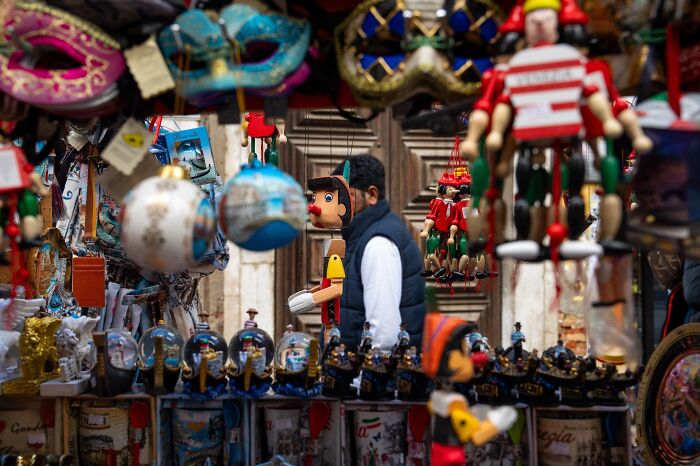 A man browses a stall filled with colorful Venetian masks and Pinocchio figures, an authentic moment of street photography.