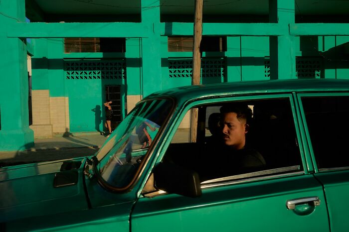 Man driving a turquoise car with a turquoise building in the background, showcasing vibrant street photography.