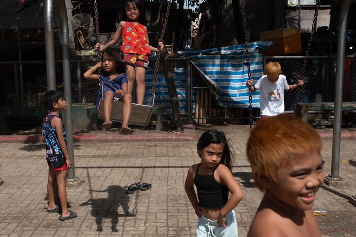 Children playing on swings and standing in a sunny outdoor area, capturing authentic moments of street photography.
