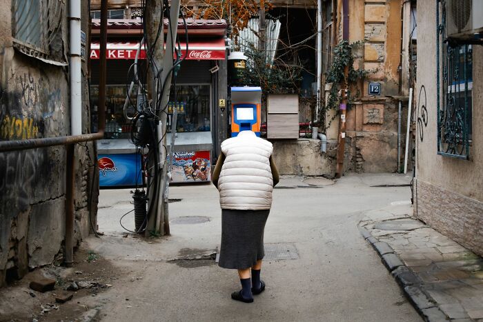 Person with a screen for a head walks down a narrow street in an urban setting, a unique street photography moment.