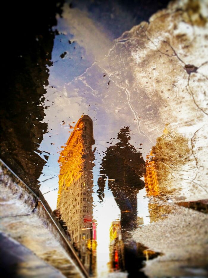 Reflection of a building and a person in a puddle, a unique perspective of street photography awards.